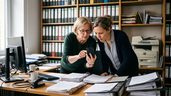 Zwei Kolleginnen prüfen gemeinsam eine E-Mail auf einem Handydisplay, umgeben von Papierstapeln und Aktenregalen