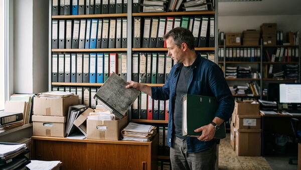 Mann zieht in einem Betriebsbüro einen Ordner aus einem deckenhohen Aktenregal, davor gestapelte Archivkartons