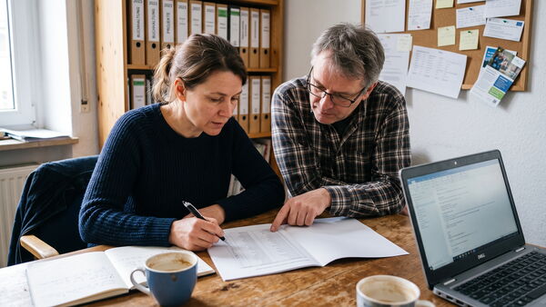 Zwei Personen gehen an einem Bürotisch gemeinsam eine ausgedruckte Namensliste durch und haken Einträge ab