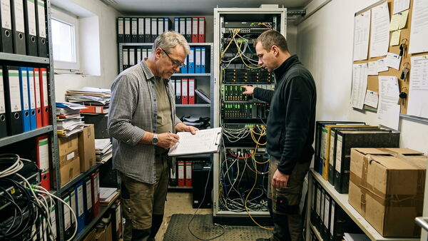Zwei Männer im Technikraum eines Büros vor einem verkabelten Serverschrank, einer macht sich Notizen auf einem Klemmbrett