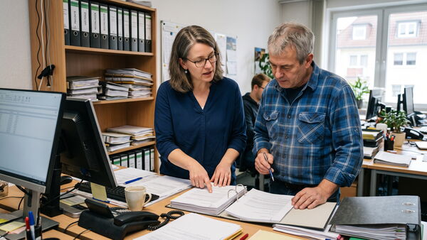 Zwei Mitarbeiter im Büro besprechen an einem Bildschirm einen Vertragsauszug, daneben ein Ordner mit Unterlagen